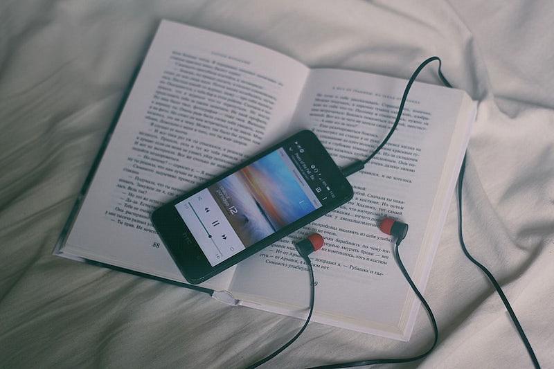 Image of open book on sheet with a mobile phone on top playing an audiobook with headphones plugged in