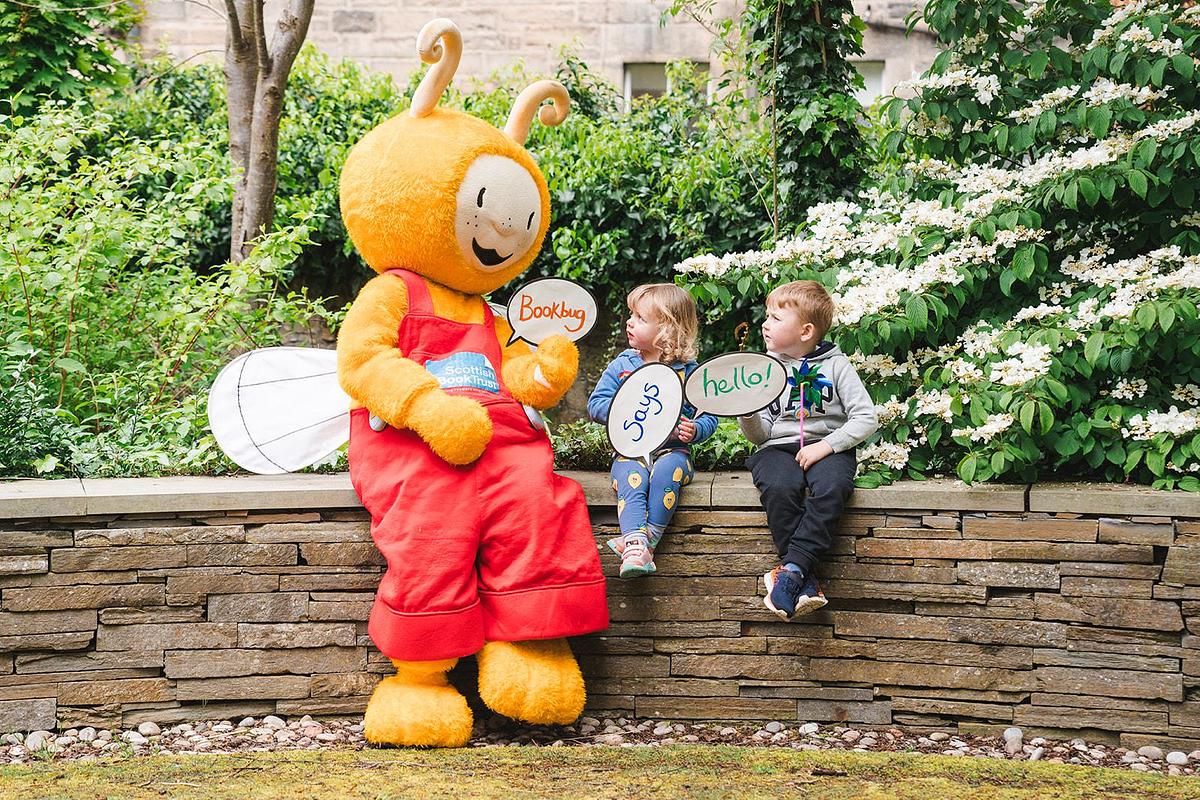 Bookbug sits on wall next to two children holding up signs saying Bookbug Says Hello!