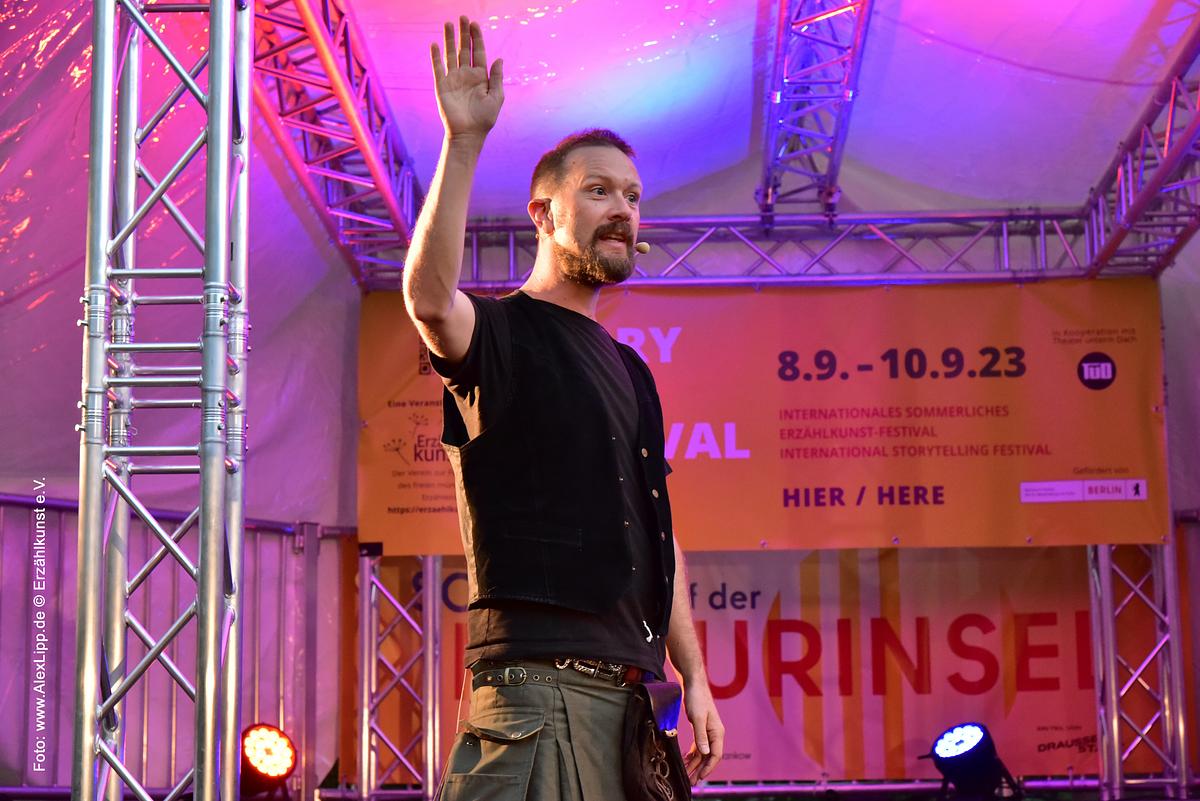 Man (Dougie Mackay) stands on stage in festival tent with arm raised