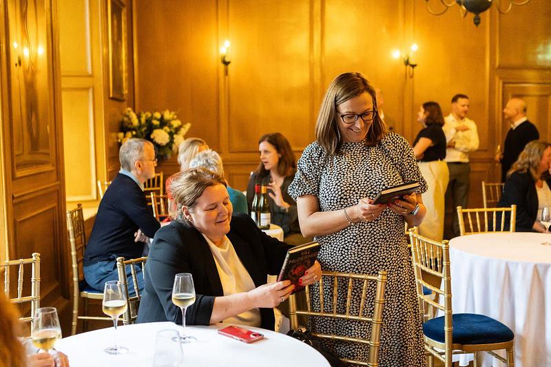 two women look at book together smiling beside a table with glasses of wine